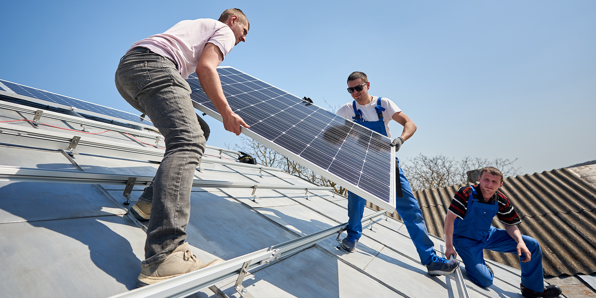 Male engineers installing solar photovoltaic panel system. Three electricians lifting blue solar module on roof of modern house. Alternative energy sustainable resources ecology concept.